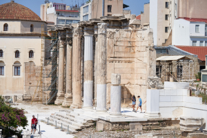 Ruinen des Tempels von Olympian Zeus in Athen, Griechenland, mit Besuchern zwischen verstreuten Steinen, Vegetation und architektonischen Fragmenten unter einem bewölkten Himmel.