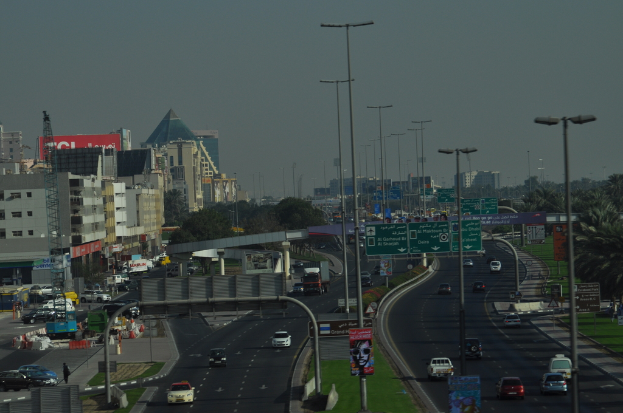 Luftaufnahme einer Stadt mit Gebäuden, Türmen, Fahrzeugen, Werbetafeln, Schildern, Straßen, Bäumen, Straßenlaternen, Menschen, Absperrgittern und Informationsanzeigen unter einem klaren Himmel.