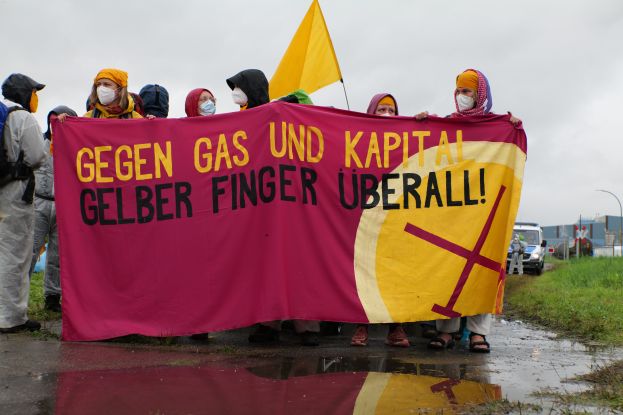 Eine Gruppe maskierter Personen mit einem Banner, auf dem 'Gegen Gas und Kapital Gelber Finger Uberall' steht, steht auf einer Straße umgeben von Gras, Fahrzeugen, Gebäuden und Laternen unter einem klaren blauen Himmel.