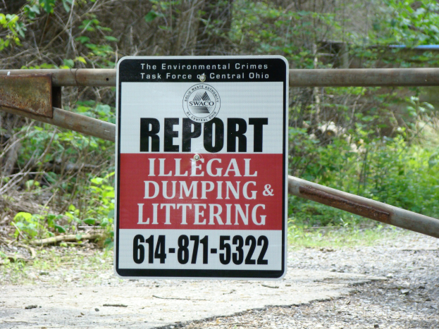 Schild am Straßenrand mit der Aufschrift "Melden Sie illegalen Müll und Littering" mit Bäumen und Pflanzen im Hintergrund und einer Tafel mit Text im Vordergrund.
