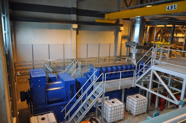 Eine Fabrikinnensicht mit Trägern, einer Treppe, Geländer, Containern, Lichtern, Rohren und Wänden.