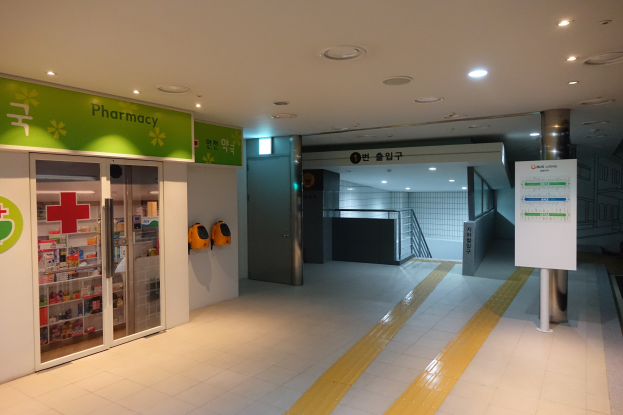 Der Eingang zu einer Apotheke mit einem "Apotheke"-Schild auf der linken Seite, einer Tafel mit Text auf der rechten Seite und beleuchtet von Lampen auf dem Dach.