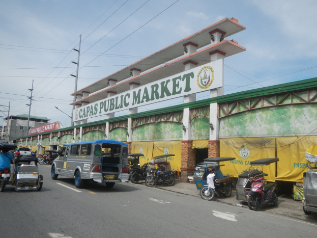 Eine belebte Stadtstraße mit Autos, Motorrädern und Rikschas, die an einem Gebäude mit der Aufschrift "Capas Public Market" vorbeifahren, vor einem Hintergrund aus Strommasten, Gebäuden und einem bewölkten Himmel.