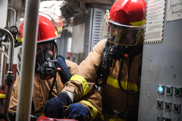 Zwei Feuerwehrleute in Schutzausrüstung, Helmen und Gasmasken arbeiten an einem Feuerwehrauto mit einer Tafel und Metallstangen im Hintergrund.