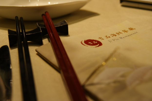Ein Tisch mit einer Speisekarte, die "Ji Pin Restaurant" auf Mandarin und Englisch lautet, zwei Paare Stäbchen (eines rot, eines schwarz) und eine weiße Schüssel vor den Stäbchen.