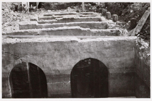 Ein Schwarz-Weiß-Foto einer einstürzenden Steinbrücke mit Bögen, umgeben von Felsen und Holzabfällen.