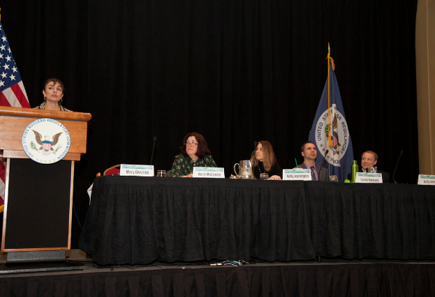 Eine Frau steht an einem Pult auf einer B├╝hne und spricht in ein Mikrofon. Vor ihr befindet sich eine runde Platte. Im Hintergrund sitzen mehrere Personen an einem Tisch mit Mikrofonen, kleinen Platten, einer Flasche, einem Glas und einem Krug. Flaggen, ein Vorhang und eine Wand sind im Hintergrund zu sehen.