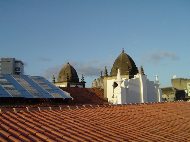 Stadtpanorama mit Gebäuden im Vordergrund und einem blauen Himmel im Hintergrund, das Solarpanels auf einem Dach zeigt.