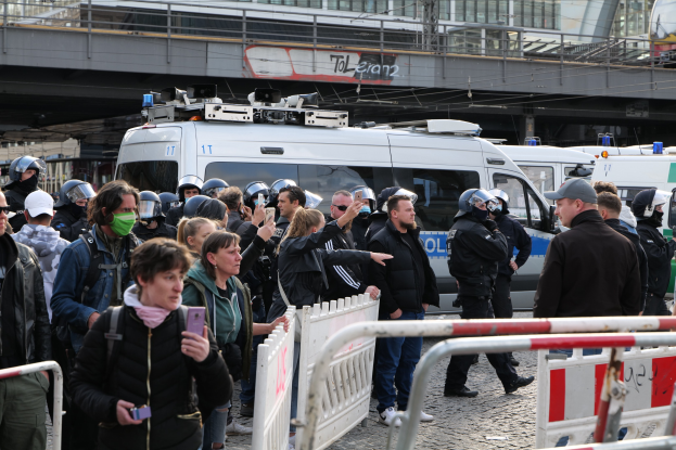 Eine Gruppe von Menschen steht vor Polizeifahrzeugen, einige tragen Helme und halten Telefone, mit Barrieren im Vordergrund und einer Brücke und Gebäuden im Hintergrund während einer Demonstration in Berlin, Deutschland.