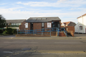 Ein Schulgebäude mit einem blauen Geländer, umgeben von Häusern, Treppen, Geländern, Texttafeln, Pflanzen, Bäumen und einem bewölkten Himmel.