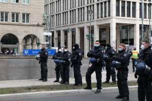 Eine Gruppe von Polizisten in schwarzen Uniformen und Masken steht vor einem Gebäude mit Glasfenstern und Säulen in Berlin, Deutschland, einige halten Helme, mit Laternen, Verkehrszeichen, Texttafeln, einer Statue, anderen Menschen und einem grasbewachsenen Boden im Hintergrund.