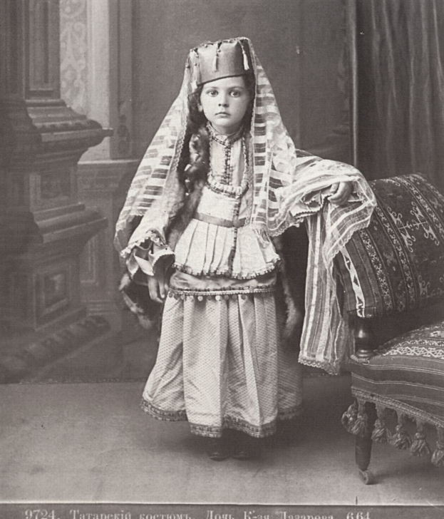 Schwarzes und weißes Foto eines jungen Mädchens in traditioneller russischer Kleidung, das neben einem Stuhl mit einem Vorhang und einer Wand im Hintergrund steht, mit Text am unteren Rand.