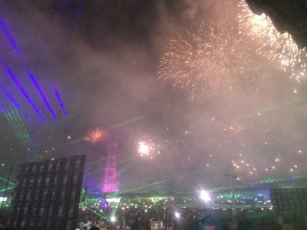 Eine Menschenmenge bei einem Konzert mit beleuchteten Gebäuden und Pfählen im Hintergrund, unter einem Himmel voller bunter Feuerwerke.