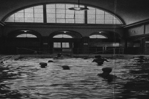 Schwarze und weiße Fotografie von Menschen, die in einem Hallenbad mit Glaswänden und einer Deckenbeleuchtung schwimmen.