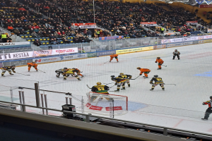 Gruppe von Menschen, die Hockey auf einer Indoor-Eisbahn mit einem Netz spielen, umgeben von einem Zaun, unter Stadionbeleuchtung, mit Zuschauern im Hintergrund und sichtbaren Bannern.