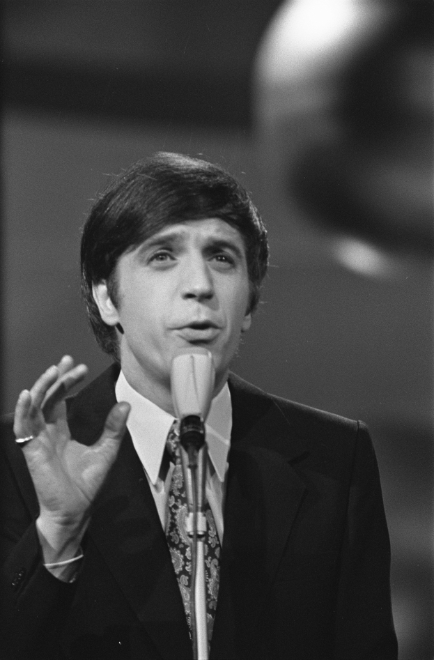 Ein Mann in einem schwarzen Anzug, weißem Hemd und gemustertem Schlips steht vor einem Mikrofon und spricht mit ernster Miene und zurückgekämmtem Haar vor einem unscharfen Hintergrund.