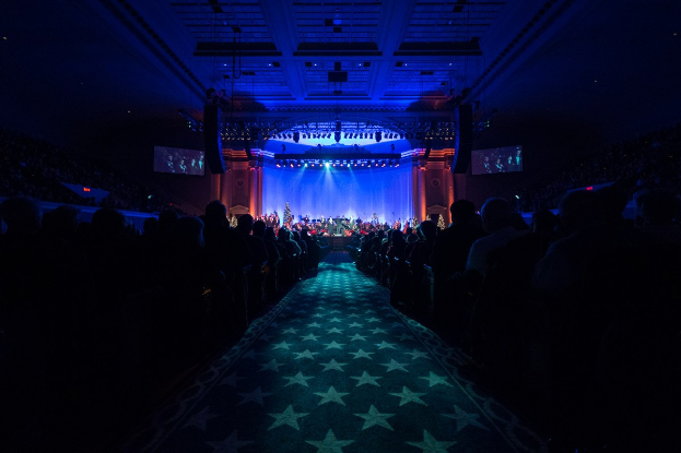 Ein großer Saal mit sitzenden Zuschauern, eine Bühne mit Musikern und Deckenbeleuchtung und -fernseher, was auf ein Konzerthaus während der Nationalkonvention 2019 hindeutet.