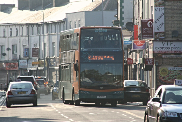 Eine Straße mit Autos und einem Bus vor Gebäuden mit Wänden, Fenstern, Tellern und Dächern, die Plakate und Banner an den Wänden zeigen, mit einem Laternenmast.