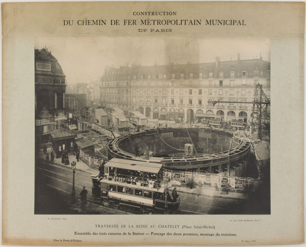 Ein altes Schwarz-Weiß-Foto einer Stadtstraße mit einem Zug auf den Schienen, umgeben von Gebäuden, Pfählen und Menschen, mit dem Text "Construction du Chemin de Fer Metropolitain Municipal de Paris" oben.