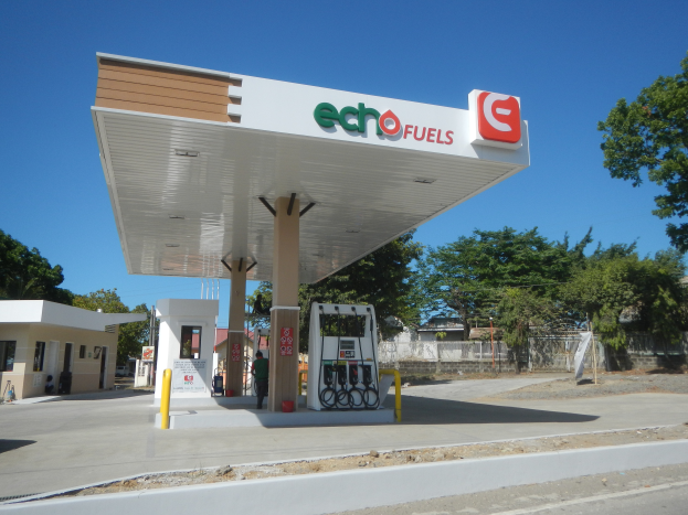Eine Tankstelle mit einem Schild, das "Echo Fuels" zeigt, im Vordergrund, umgeben von Gebäuden, Bäumen und einem Zaun, mit einer Straße unten und dem Himmel oben.