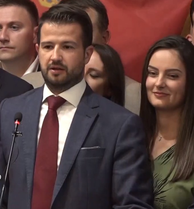 Mann in Anzug und Krawatte, der vor einem Publikum an einem Rednerpult steht und ein Mikrofon hält, mit einem Banner im Hintergrund.