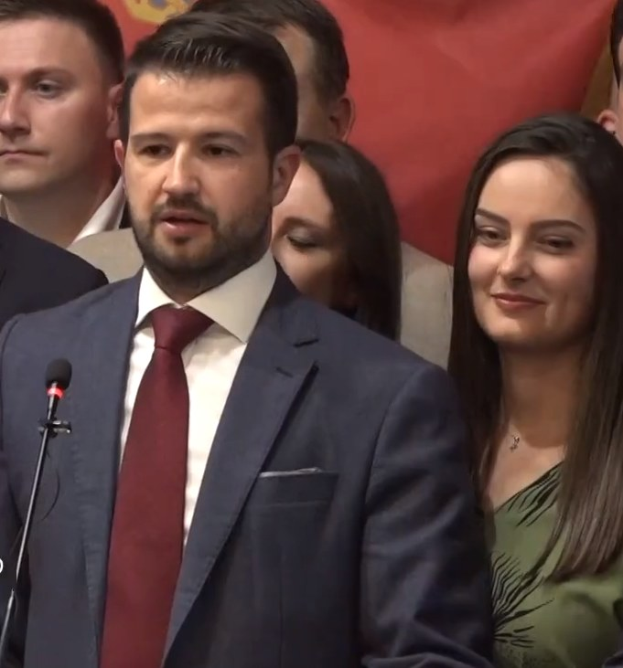 Mann in Anzug und Krawatte, der vor einem Publikum an einem Rednerpult steht und ein Mikrofon hält, mit einem Banner im Hintergrund.