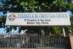 Eine evangelische christliche Kirche in Bacoor City, Cavite, mit einem Schild, einem Metallzaun, einer Wand, Bäumen, Fahrzeugen, Gebäuden, Polen und einem bewölkten Himmel.