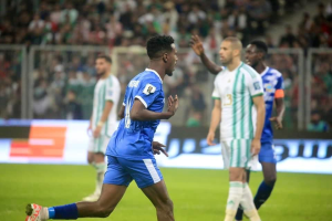 Ein Fussballspieler in einem blauen und weißen Trikot feiert ein Tor, umgeben von einer Gruppe in weißen und grünen Trikots, mit einem unscharfen Hintergrund, der ein weißes und blaues Brett und ein Geländer zeigt.