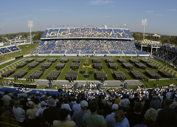 Eine große Menschenmenge schaut einem Fußballspiel in einem grasbewachsenen Stadion unter einem klaren blauen Himmel zu, mit Laternenmasten, Bäumen und Gebäuden im Hintergrund.