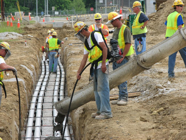 Eine Baustelle mit Arbeitern in Jacken und Helmen, Absperrgittern, ZÃ¤unung, BÃ¤umen, Masten, Erde und Rohren im Hintergrund.
