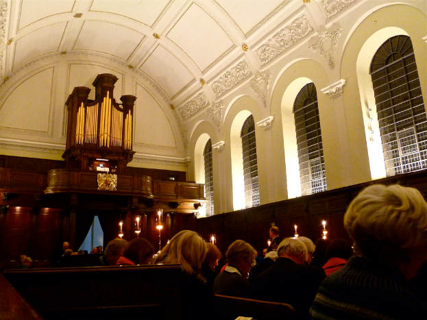 Das Innere einer Kirche mit sitzenden Männern und Frauen, Kerzen vor ihnen und weißen Wänden und Dach.