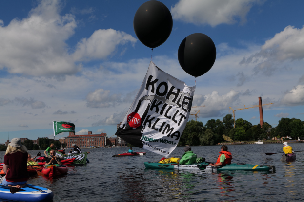 Gruppe von Menschen in Kajaks auf dem Wasser paddeln mit einem Banner, auf dem "Kohle Kill Klima" steht und Bäume, Gebäude und Kräne im Hintergrund unter einem klaren blauen Himmel.