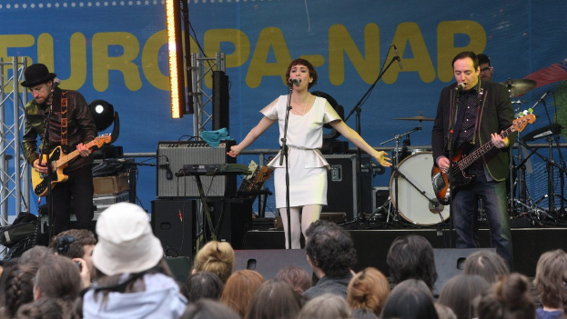 Vier Personen auf der Bühne: eine Frau in einem weißen Kleid, die singt, zwei Männer, die Gitarren spielen, und eine Menge unten, mit einer Wand und Lautsprechern im Hintergrund.