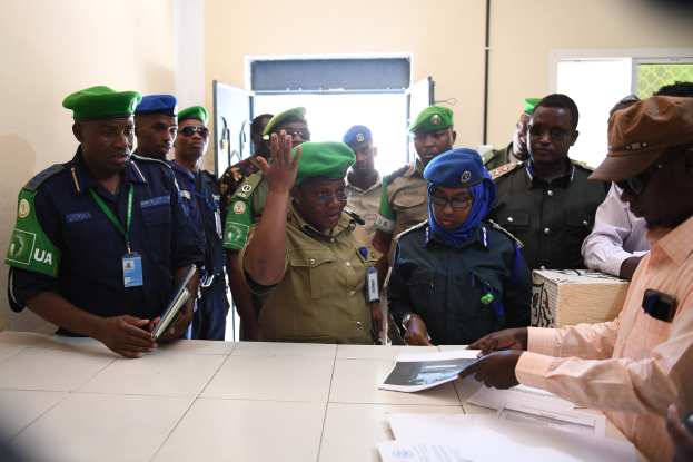 Eine Gruppe von Menschen steht um einen Tisch mit Papieren, einige tragen Mützen und ID-Karten, einer hält ein Mobiltelefon, in einem Raum mit Türen und Fenstern.