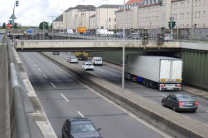 Eine Autobahn mit Autos und Lastwagen, eine Brücke im Hintergrund, Laternenpfähle, Verkehrsampeln, Gebäude mit Fenstern, Bäume und ein bewölkter Himmel.