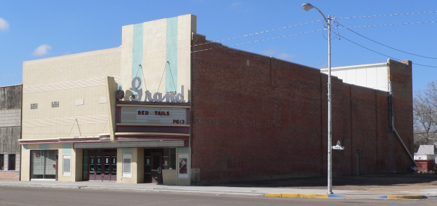 Alte Kinokasse an einer Straßenecke mit umliegenden Gebäuden, Straßeninfrastruktur, Fahrzeugen, Bäumen und einem bewölkten Himmel.