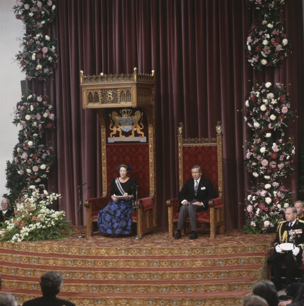 Königin Elizabeth II und Prinz Philip, Herzog von Edinburgh, sitzen vor einem Publikum, wobei die Königin ein blaues Kleid trägt und ein Buch hält, und der Herzog eine schwarze Blazer, ein weißes Hemd und schwarze Hosen trägt, vor einem maronenfarbenen Vorhang mit Blumenbouquets auf beiden Seiten.