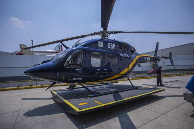 Polizeihubschrauber auf dem Flughafenparkplatz mit einer Person davor, Flugzeuge und ein Auto im Hintergrund unter einem klaren blauen Himmel.