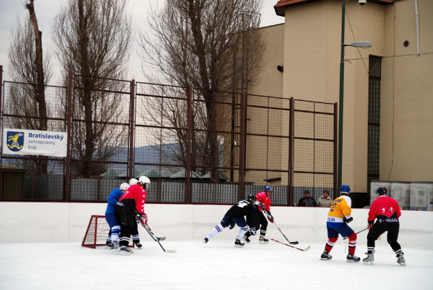 Menschen beim Eislaufen auf einer Eisbahn mit einem Hintergrund aus Himmel, Bäumen, Gebäuden, einer Straßenlaterne, einem Namensschild und Zäunen.
