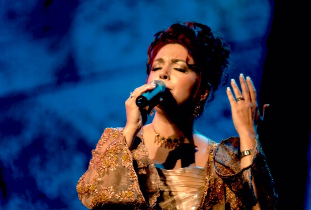 Frau in goldenem Kleid, die in ein Mikrofon singt, auf einer Bühne beim Eurovision Song Contest mit blau-schwarzem Hintergrund.