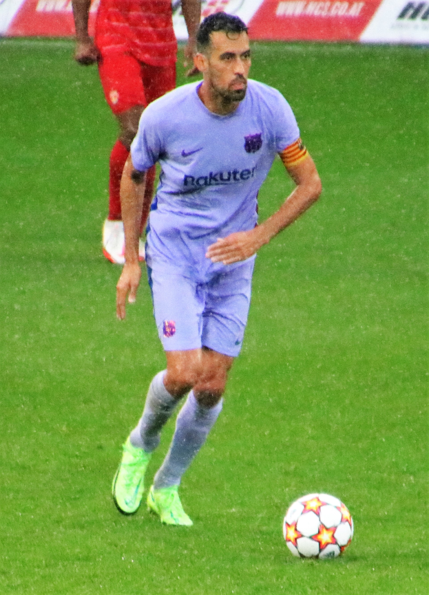 Ein Fußballspieler in einem Sportoutfit und -schuhen ist im Einsatz auf dem Feld, wobei eine Tafel mit Text im Hintergrund sichtbar ist.