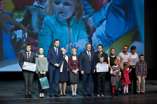 Eine Gruppe von Menschen auf einer Bühne, einige halten Taschen und Urkunden, im Hintergrund ein Bildschirm mit einem Kind und anderen Menschen.