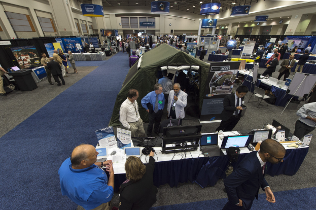 Eine große Gruppe von Menschen auf einer Karriere-Messe, die um einen Tisch mit Laptops, Monitoren und Papieren stehen, mit einem Zelt, Schildern, Deckenleuchten und Fenstern im Hintergrund.