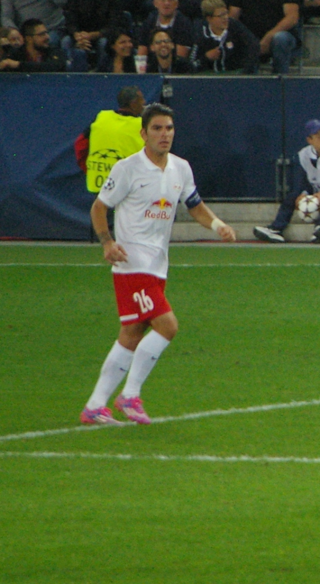 Mann in weißem Hemd und roten Shorts spielt Fußball auf einem Feld, im Hintergrund sitzen Menschen.
