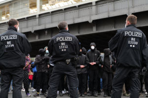 Polizisten in schwarzer Uniform und Masken vor einer Menge während einer Demonstration, mit einer Brücke und einem Gebäude im Hintergrund.