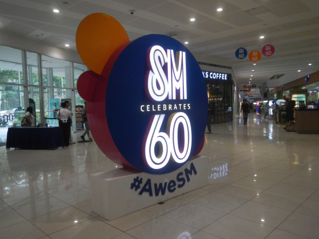 Eine Gruppe von Menschen steht auf einem Einkaufszentrumsboden unter einem 'SM feiert 60-jähriges Jubiläum'-Schild, mit einem schwarzen-gedeckten Tisch auf der linken Seite und Glastüren, Schildtafeln und Deckenleuchten im Hintergrund.