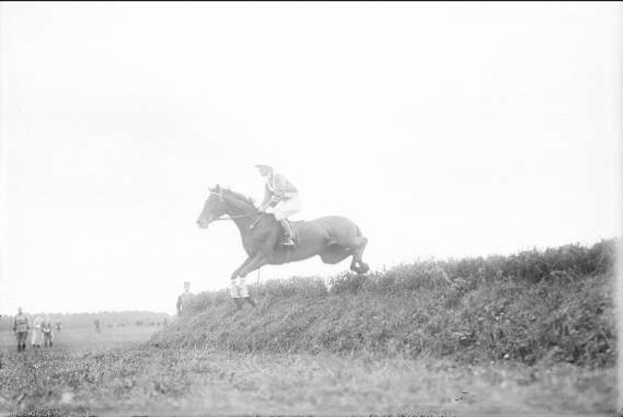 Schwarze und weiße Fotografie eines Pferdes und Reiters, die über einen grasbewachsenen Hügel springen, wobei der Reiter einen Helm und einen langen Mantel trägt und Zuschauer im Hintergrund zu sehen sind.