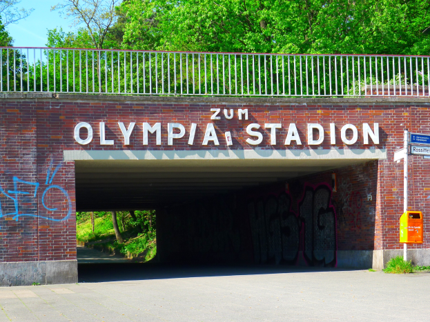 Der Eingang zum Olympiastadion in Berlin, Deutschland, mit einer Brücke, einem Metallzaun, einem Schild, einer Box, Pflanzen, Gras, einer Baumgruppe und einem bewölkten Himmel.