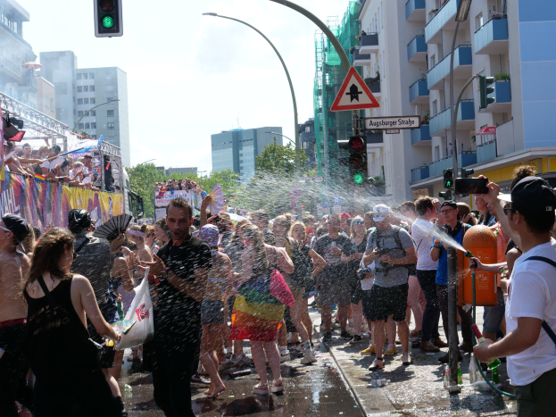 Menschen auf einer Christopher Street Day Parade, die sich gegenseitig mit Wasser bespritzen und Gegenstände halten, mit einem Banner links und Gebäuden, Bäumen und Straßen-Elementen im Hintergrund.