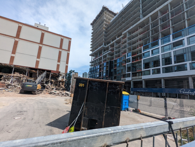 Großes Gebäude mit umfangreichem Schutt davor, umgeben von einem Metallzaun, umgeben von einem Container, Pflanzen, Bäumen, Pfählen, einem Schild, Fahrzeugen, Steinen und einem bewölkten Himmel, was auf eine Baustelle für einen neuen Wohnkomplex hinweist.